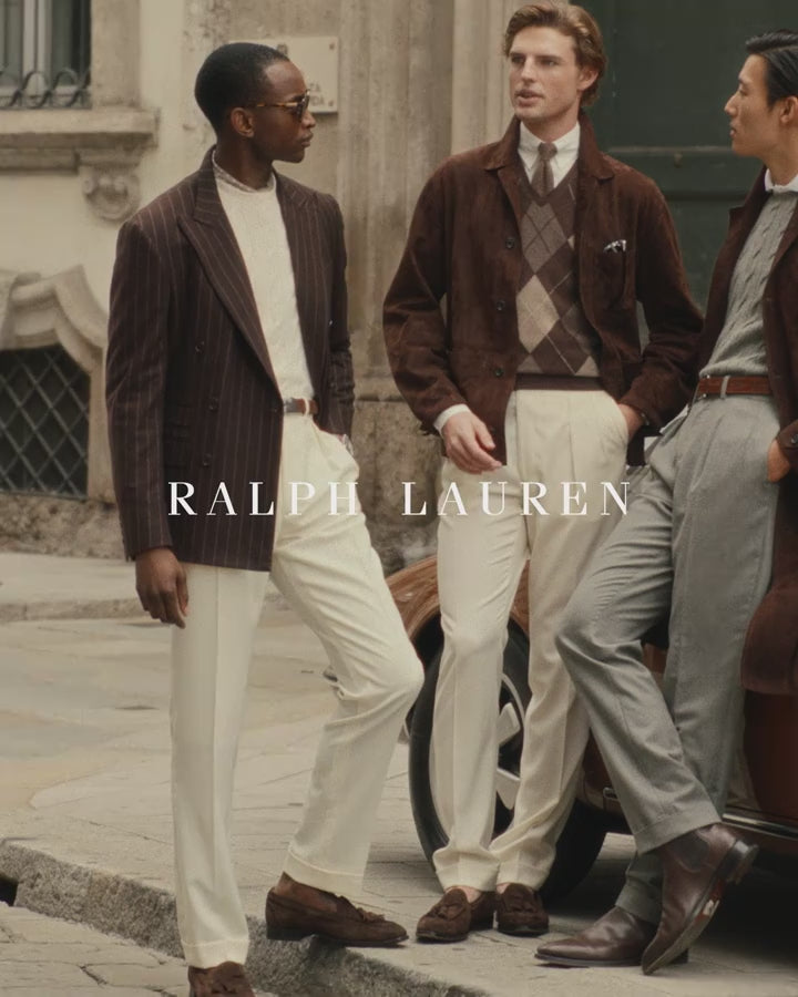 Man wearing an espresso suede jacket, ivory knit polo and ecru tailored trousers in a sunlit courtyard.